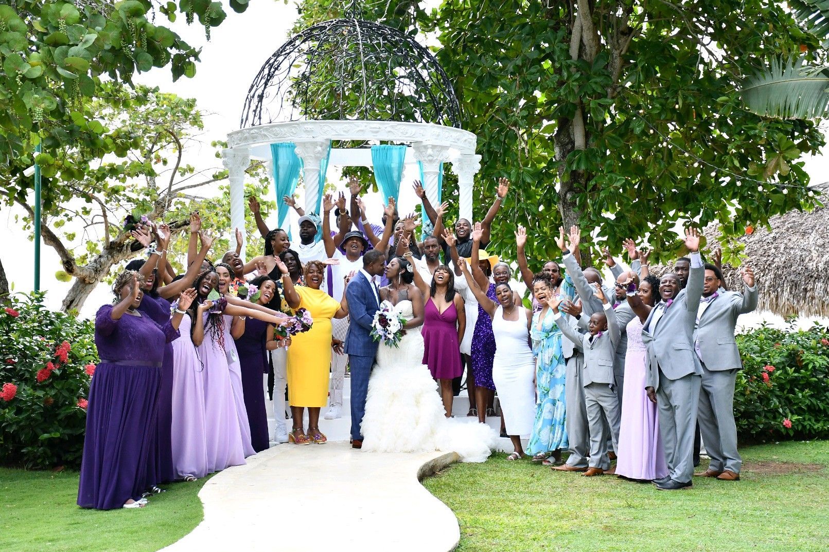 Wedding group poses with bride and groom, near a gazebo. Everyone raises hands in celebration.