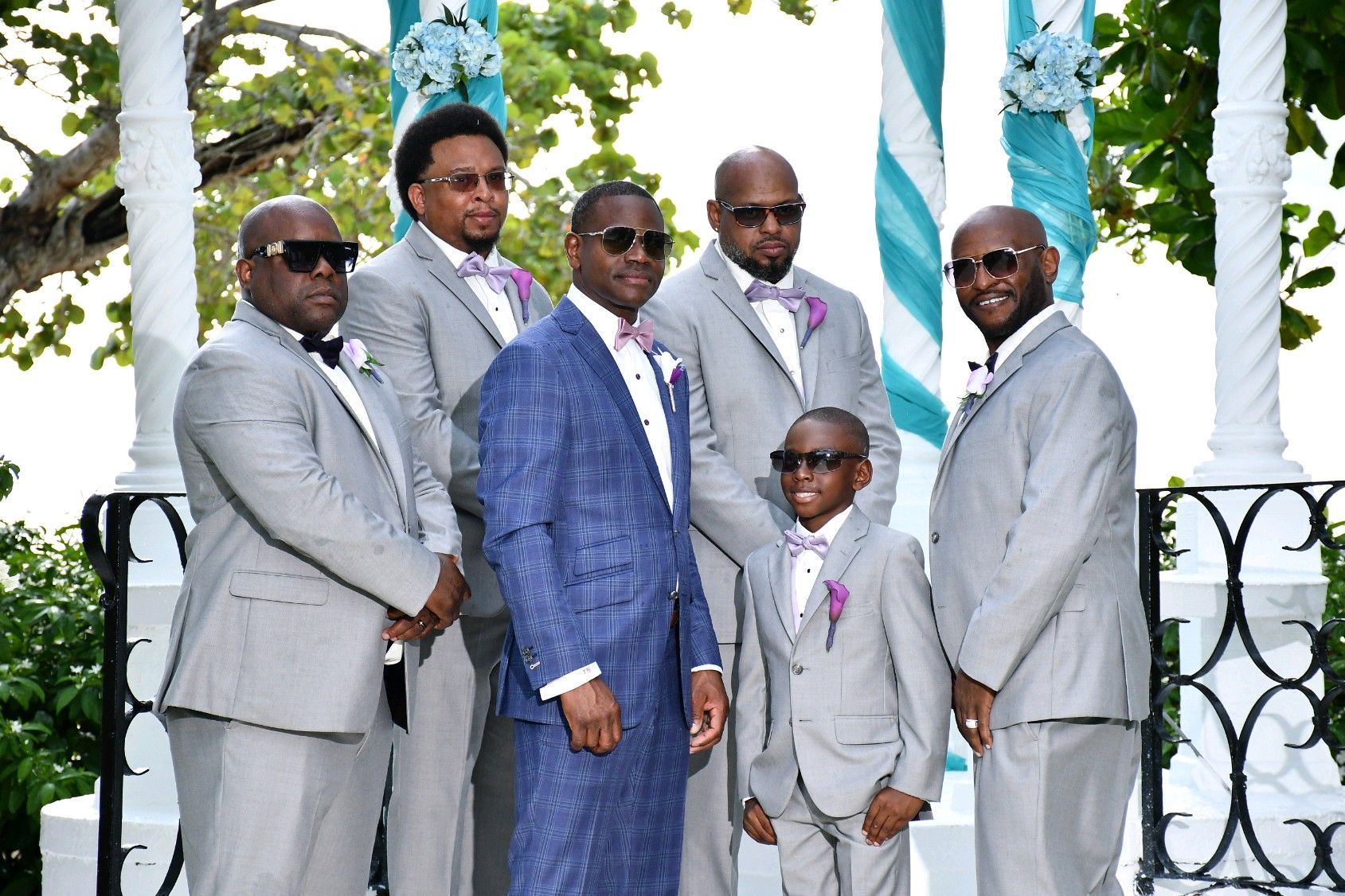 Men in formal wear posing for a photo outdoors; one in a blue suit, others in gray suits with bow ties.