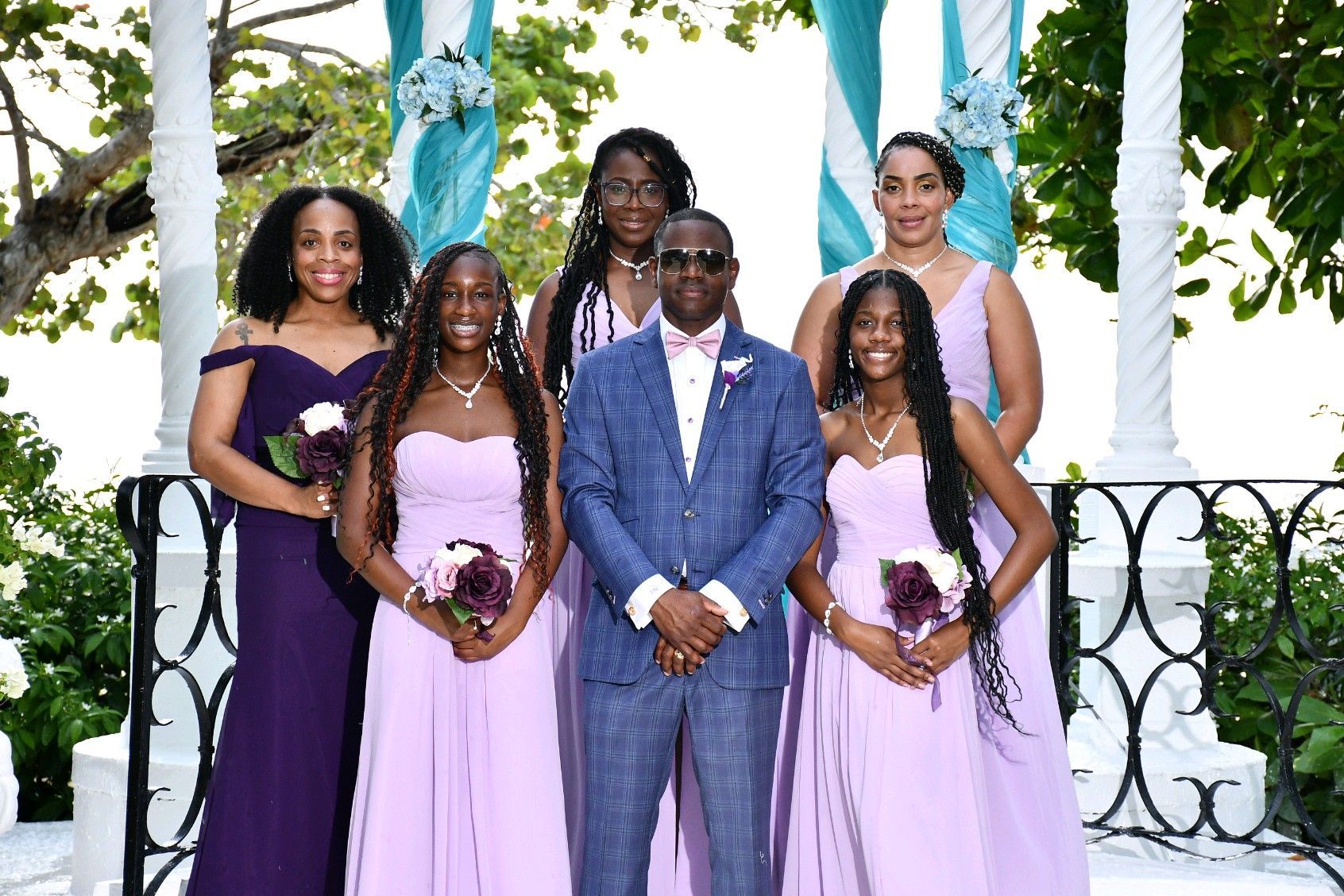 Wedding group, groom in blue suit with bridesmaids in lavender dresses, outdoor setting.
