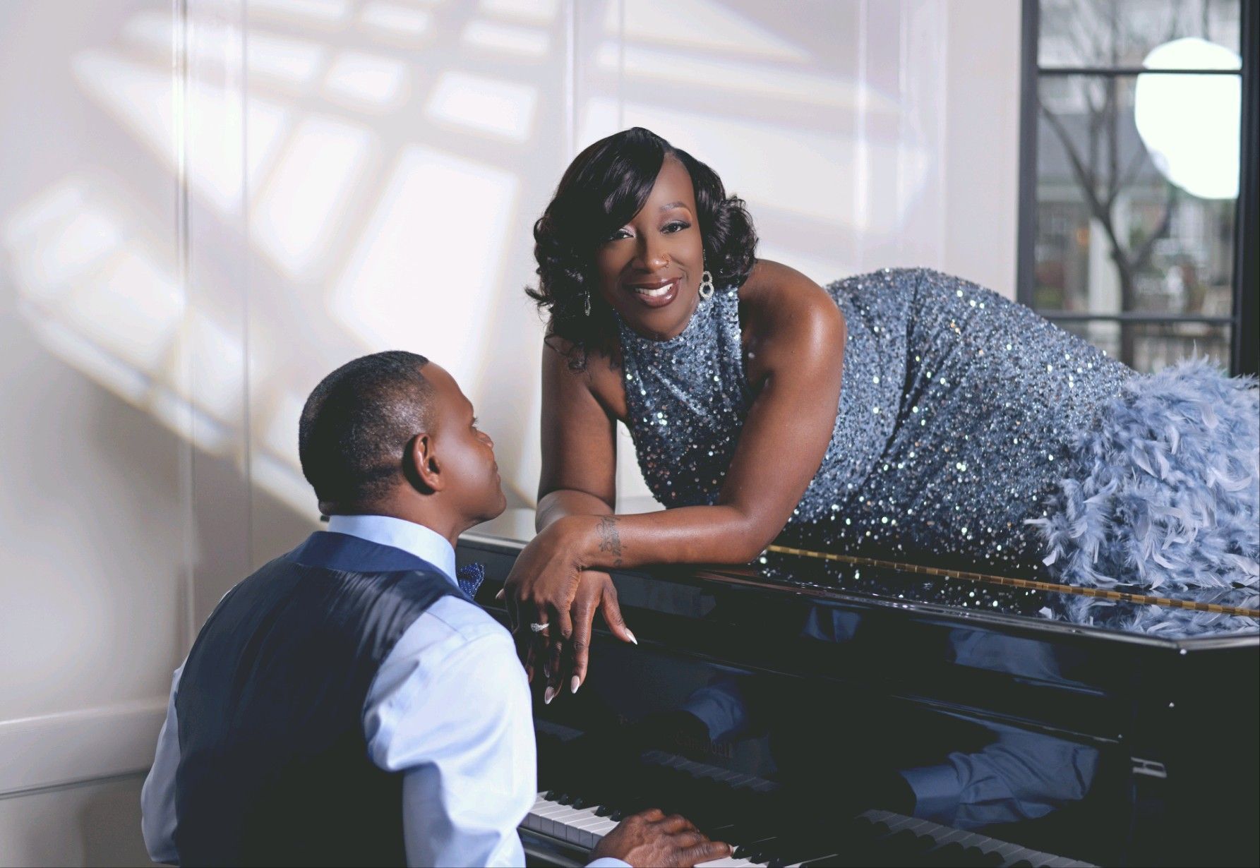 Woman in gown reclines on piano as man plays; elegant interior.
