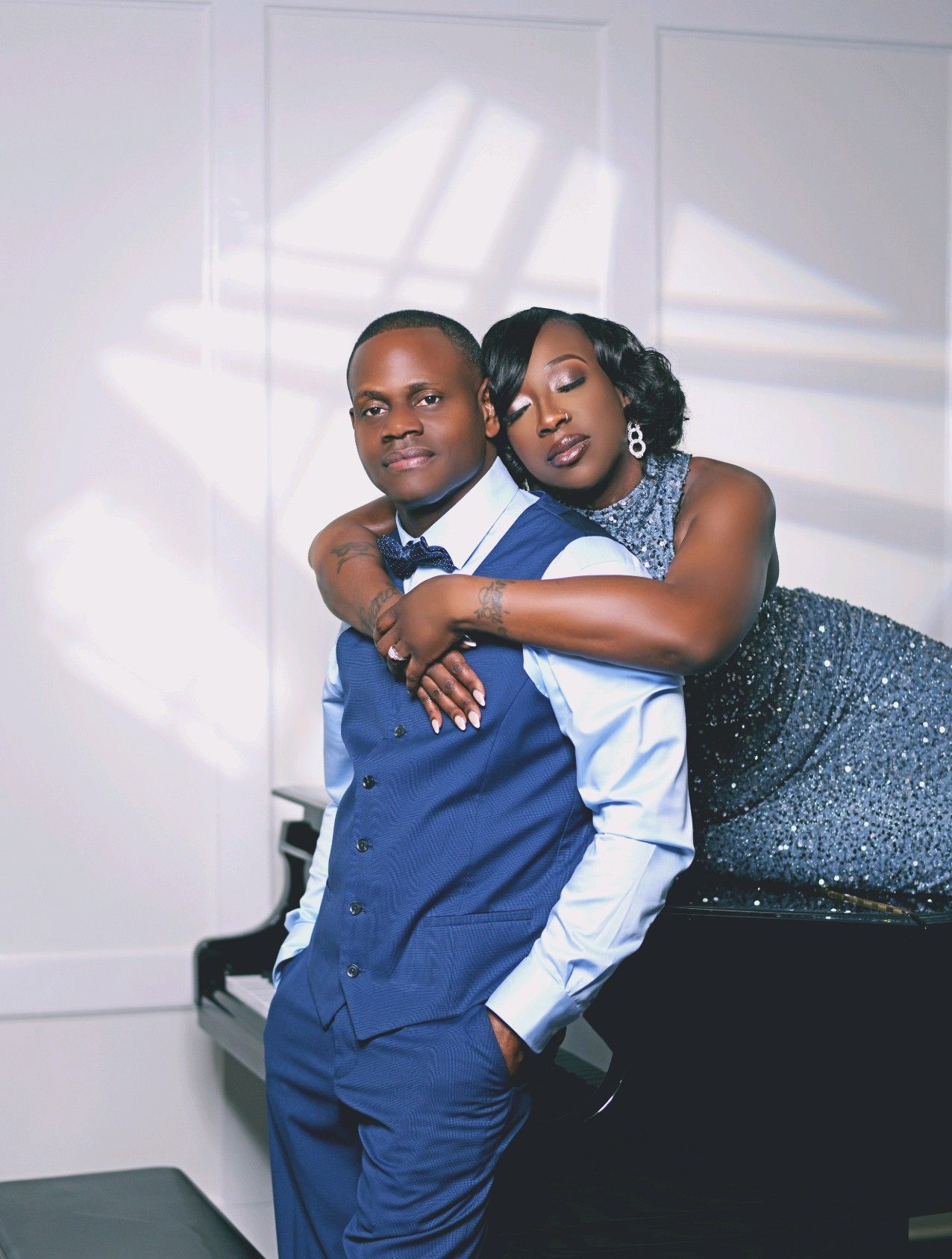 Couple in formal attire, woman leaning on man's shoulder, near a grand piano. White walls, elegant setting.