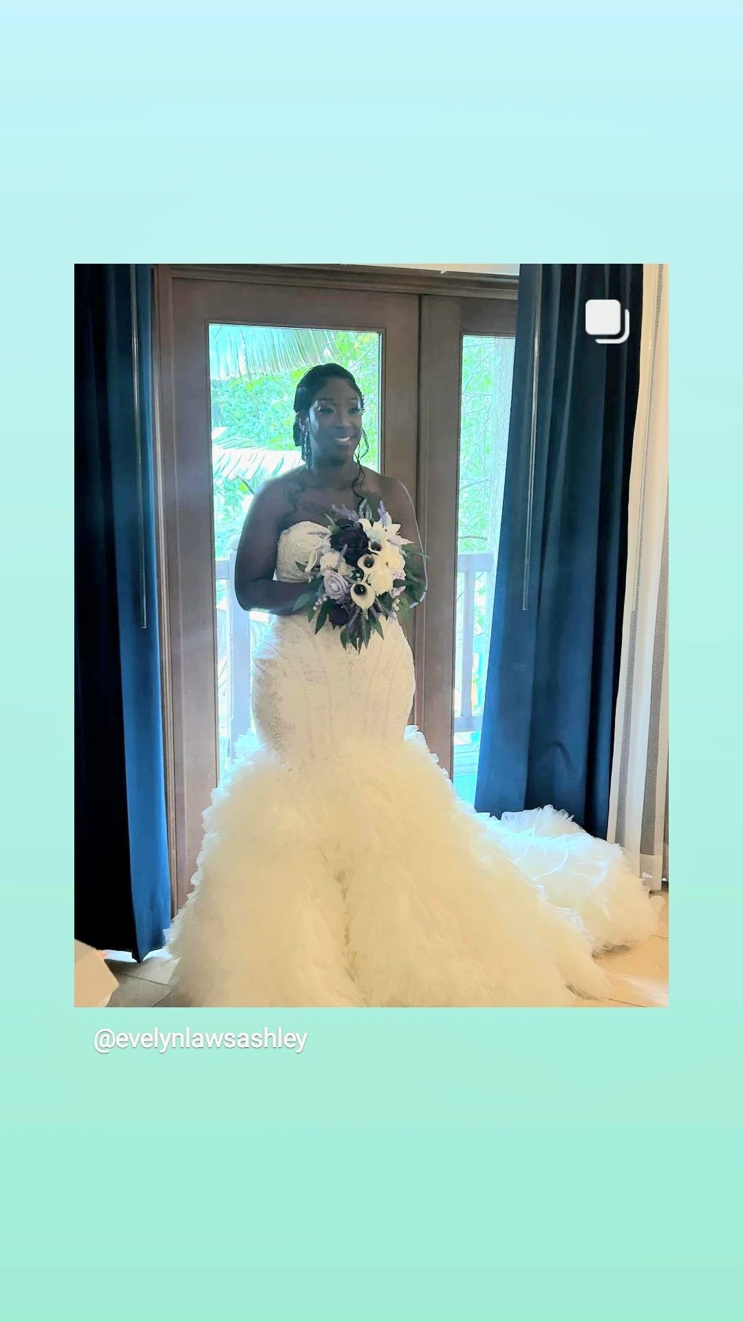 Bride in a strapless, ruffled white gown, holding a bouquet, standing by a window.