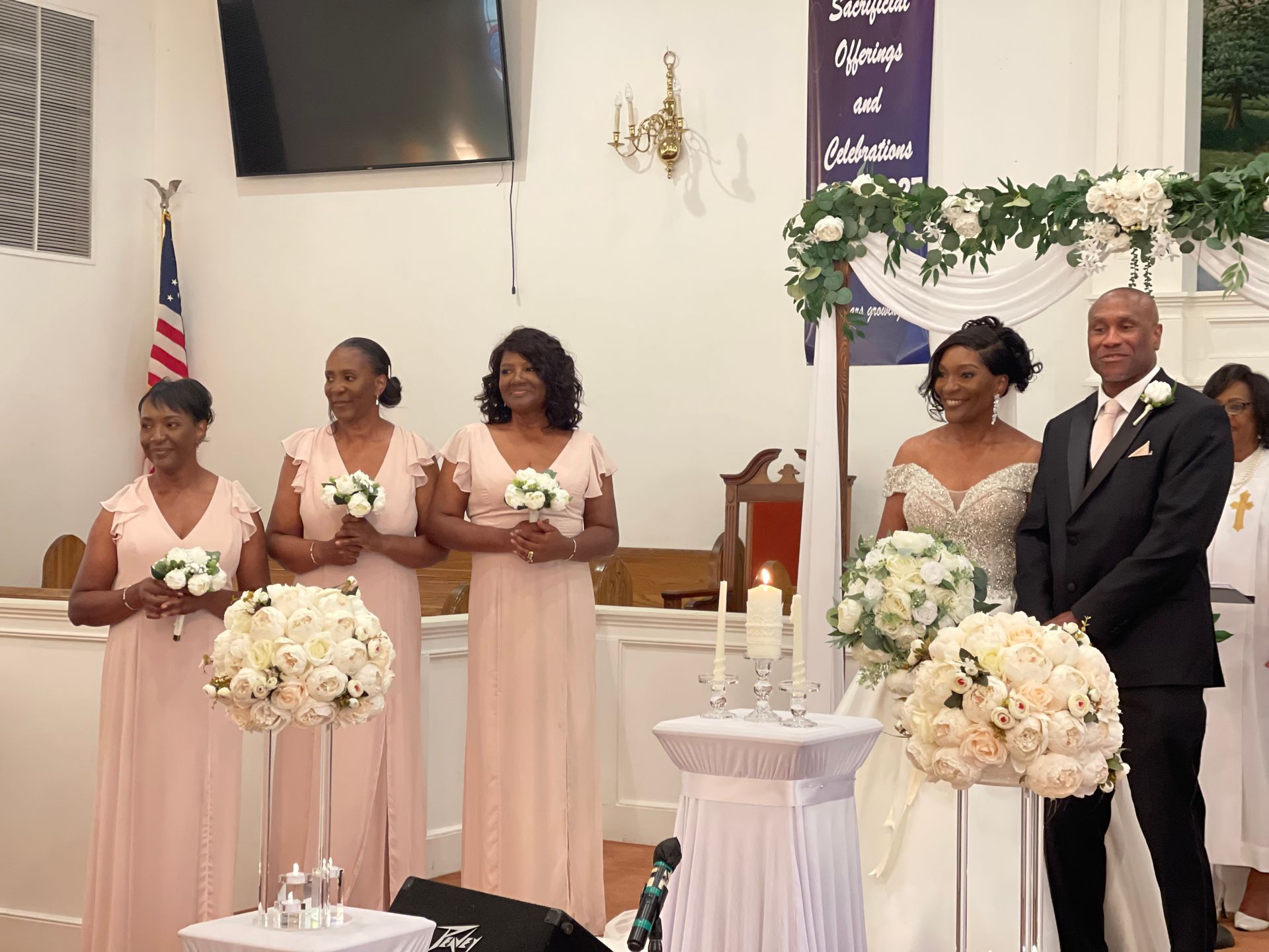 Wedding ceremony: Bride and groom with bridesmaids in a church, decorated with flowers, American flag, and arch.