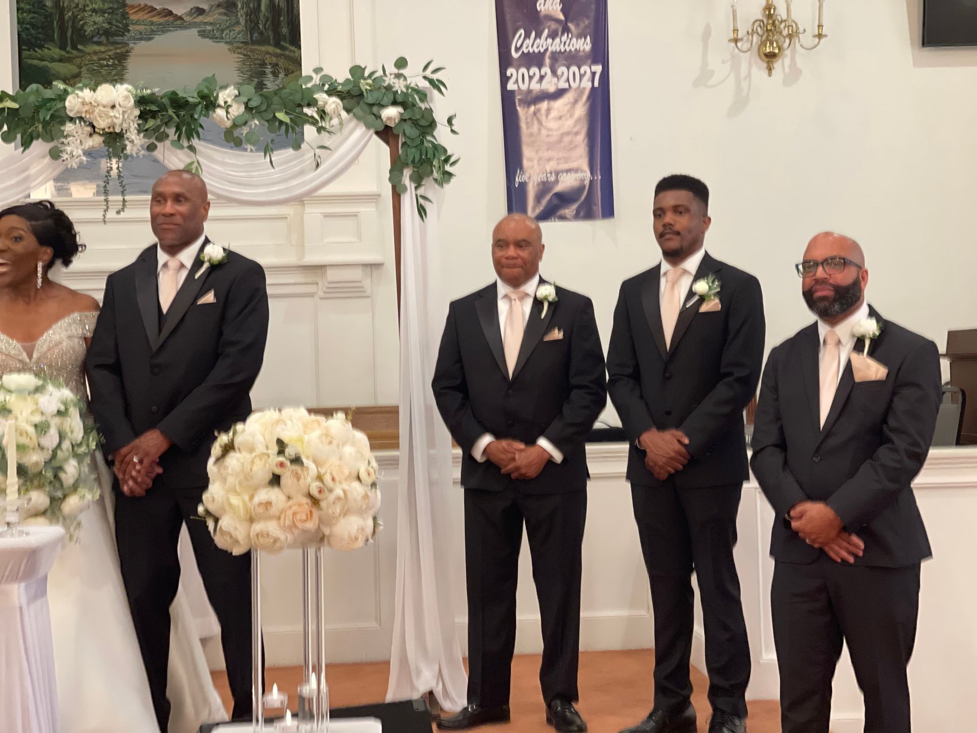 Wedding ceremony: Bride, groom, groomsmen in black suits, standing under an arch, flowers and smiles.