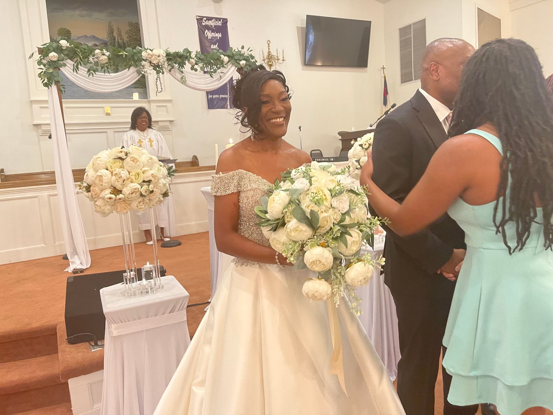 Bride smiling, holding bouquet, at wedding ceremony in church. Guests, floral decorations.