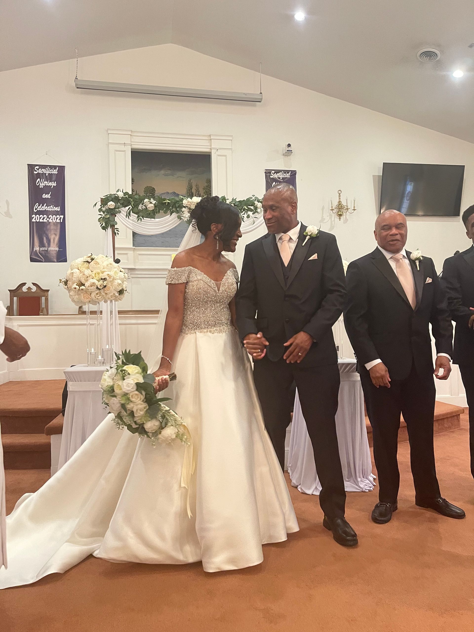 Bride and groom at the altar, smiling, holding hands. Bride in white gown, groom in black suit, church setting.