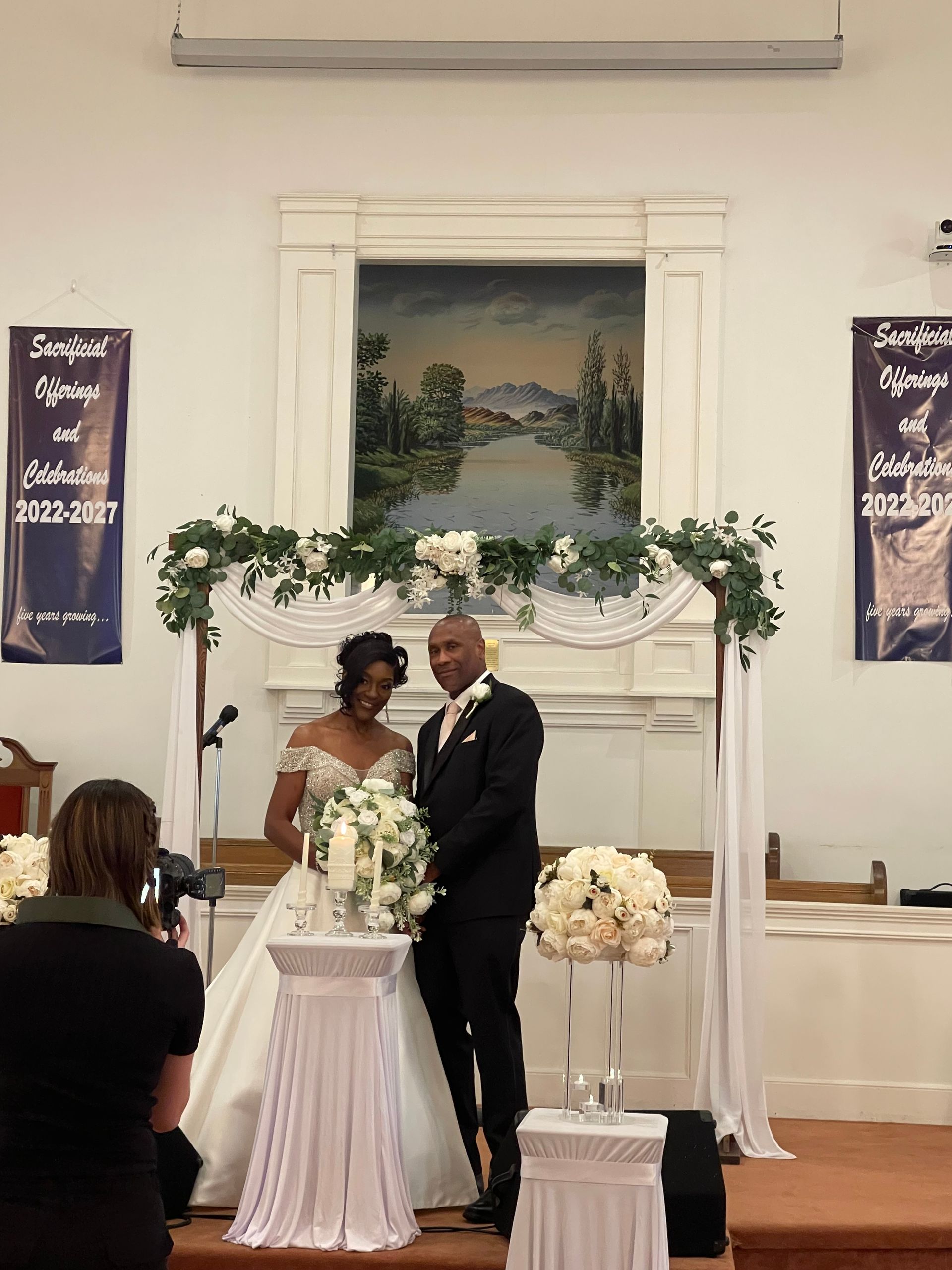 Couple in wedding attire at altar; woman in white dress, man in suit, floral arch, photographer.