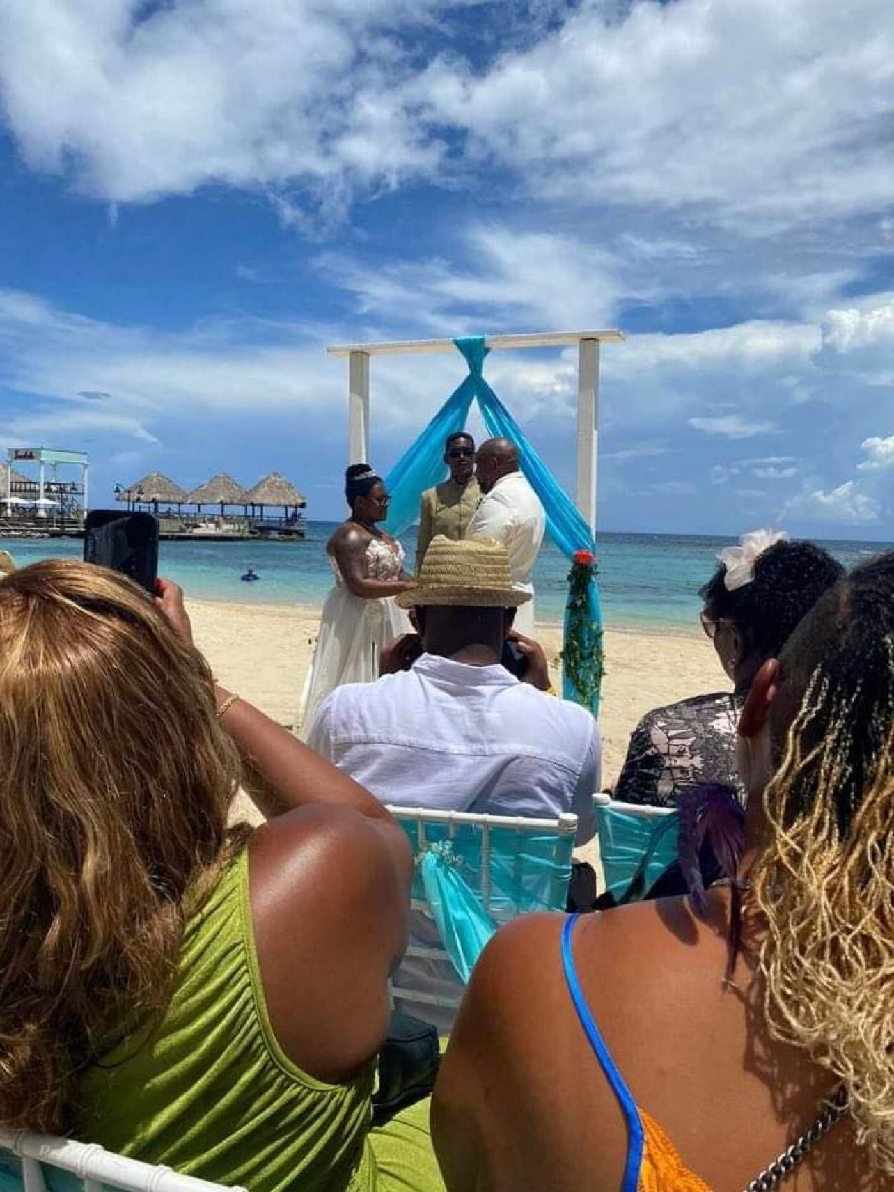 Beach wedding with bride, groom, officiant under arch; guests seated, taking photos. Blue sky, ocean.