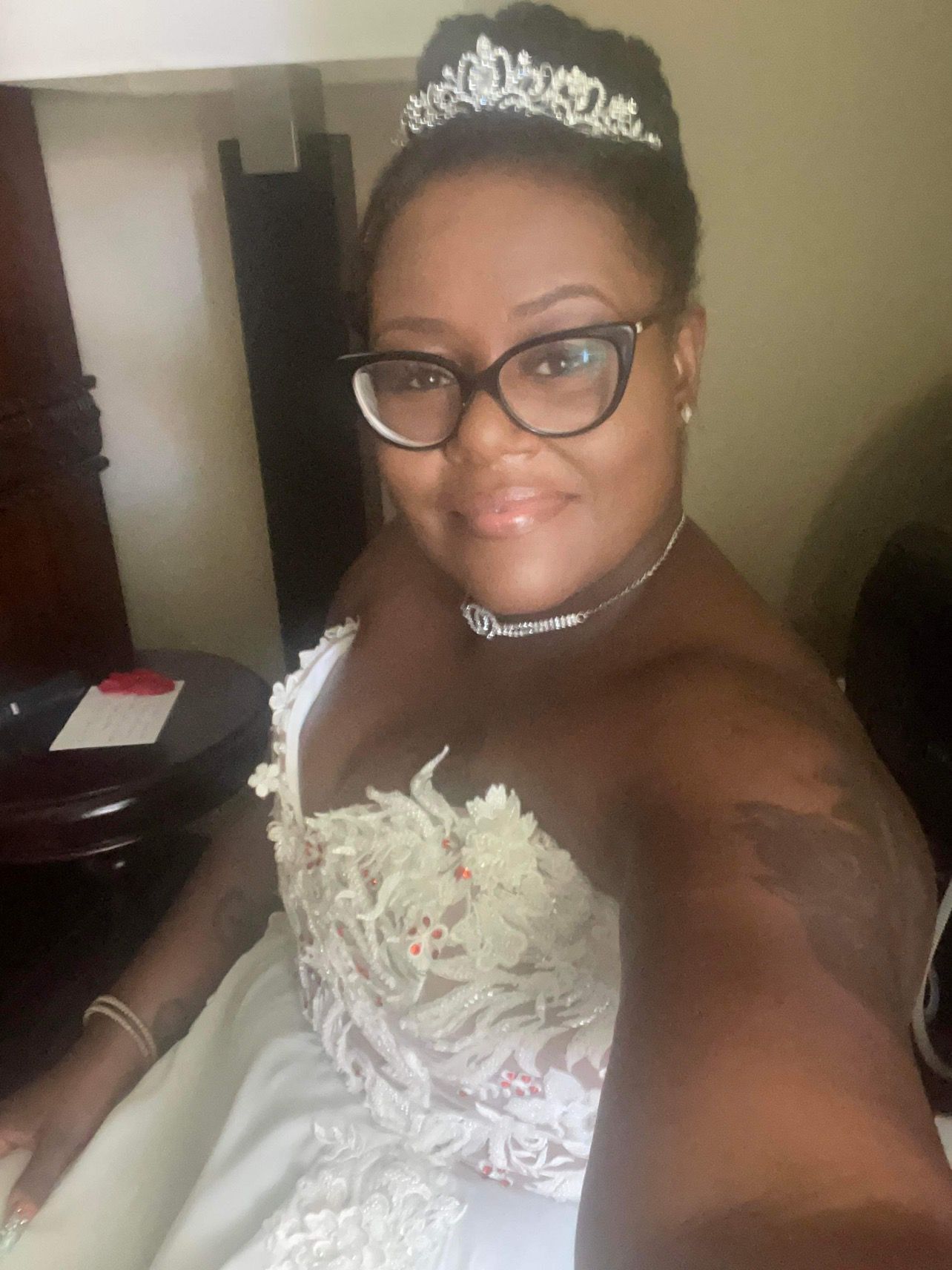 Bride wearing a wedding dress and glasses, with a tiara, sitting in a room with dark furniture.