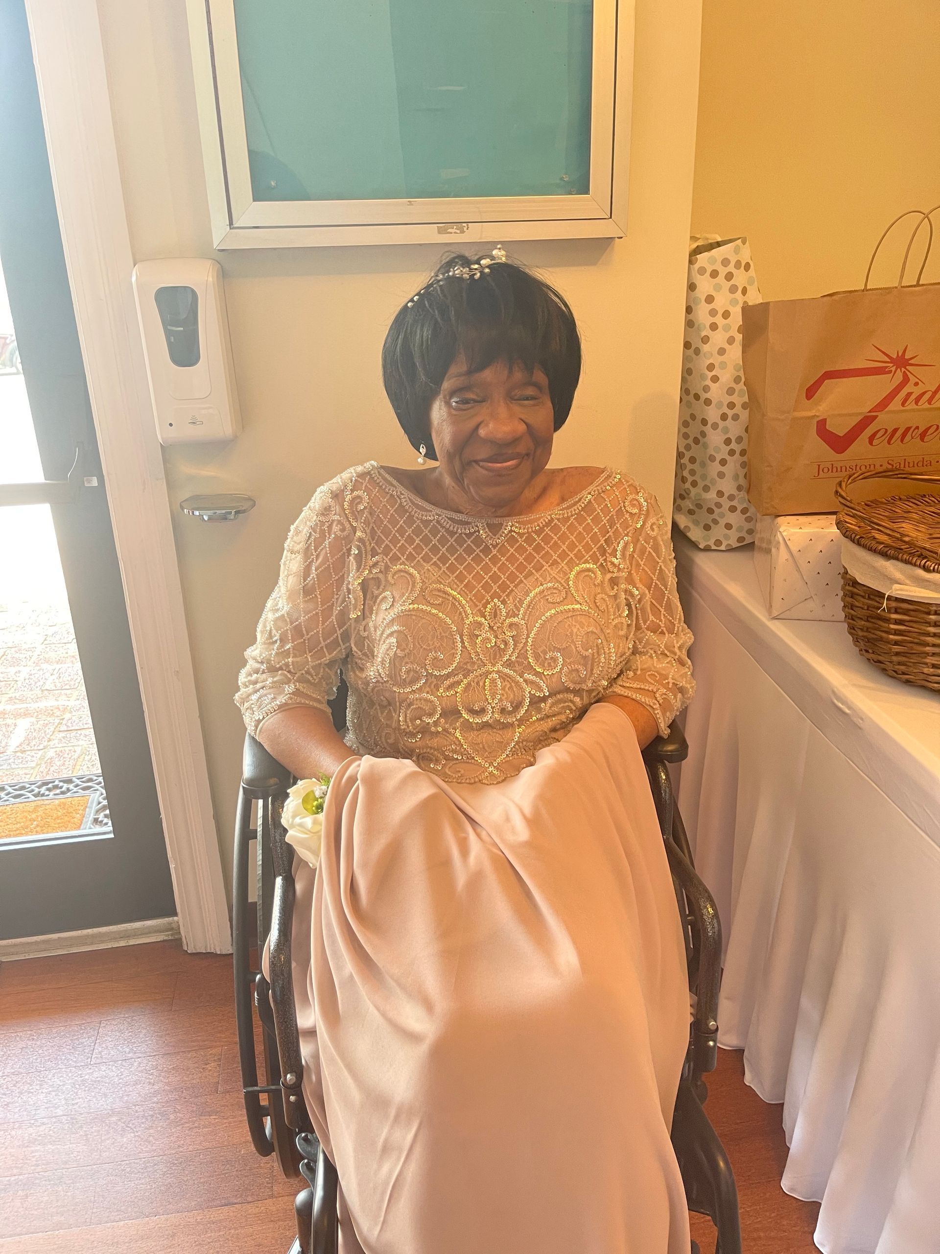 Elderly Black woman in a peach-colored gown, seated in a wheelchair. She smiles, in a well-lit room.
