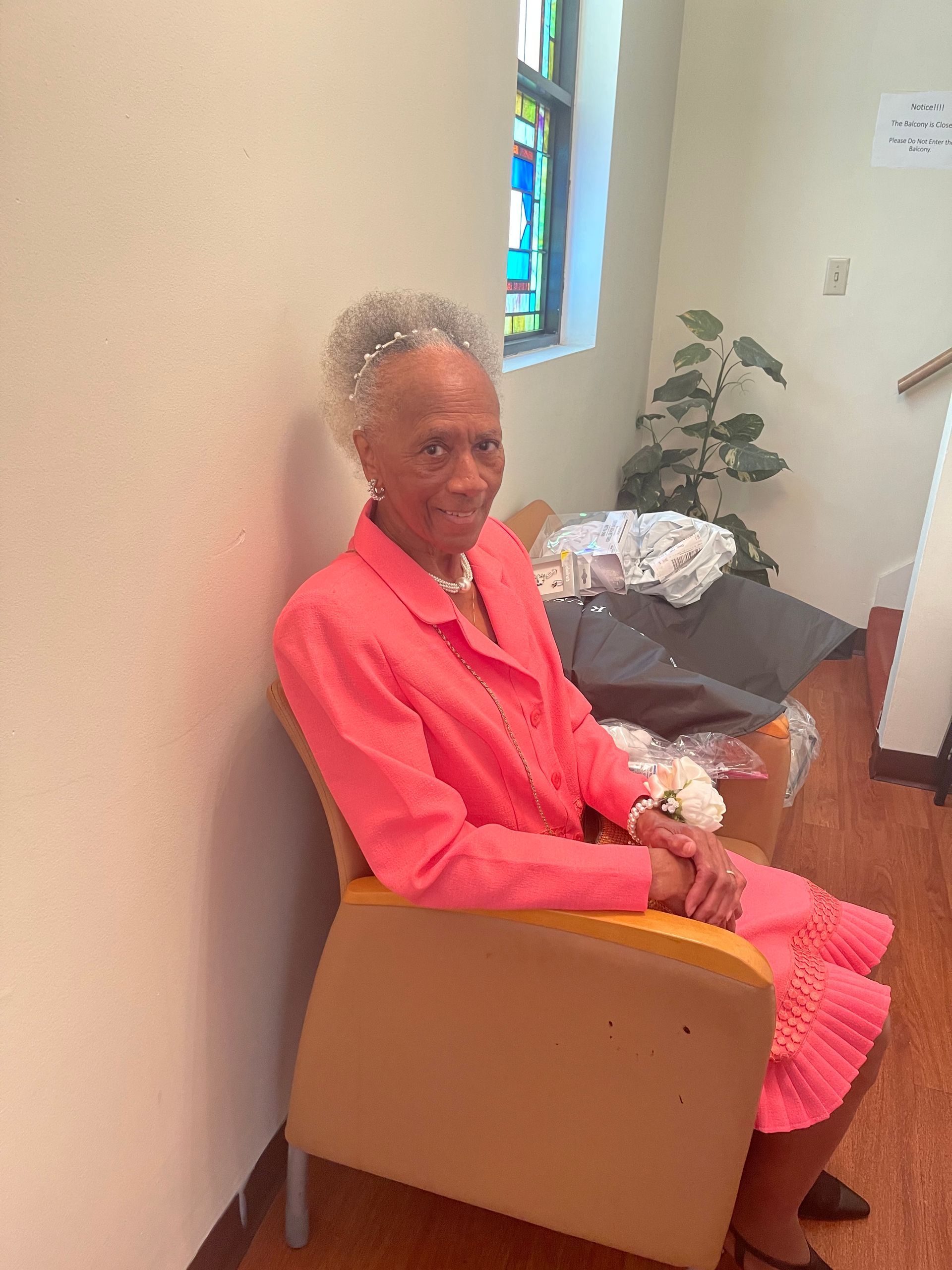 Elderly Black woman in pink suit smiles, seated in a church with stained glass.