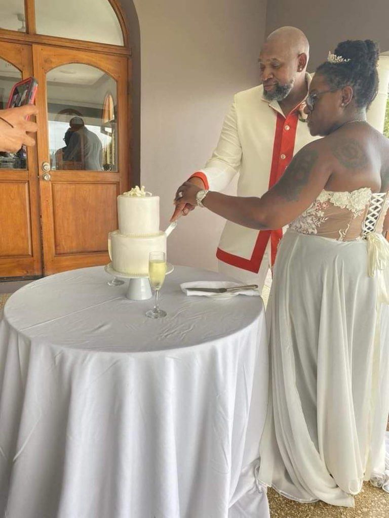 Couple cutting wedding cake at an outdoor reception. The man wears a white suit with red trim. The bride wears a white dress.