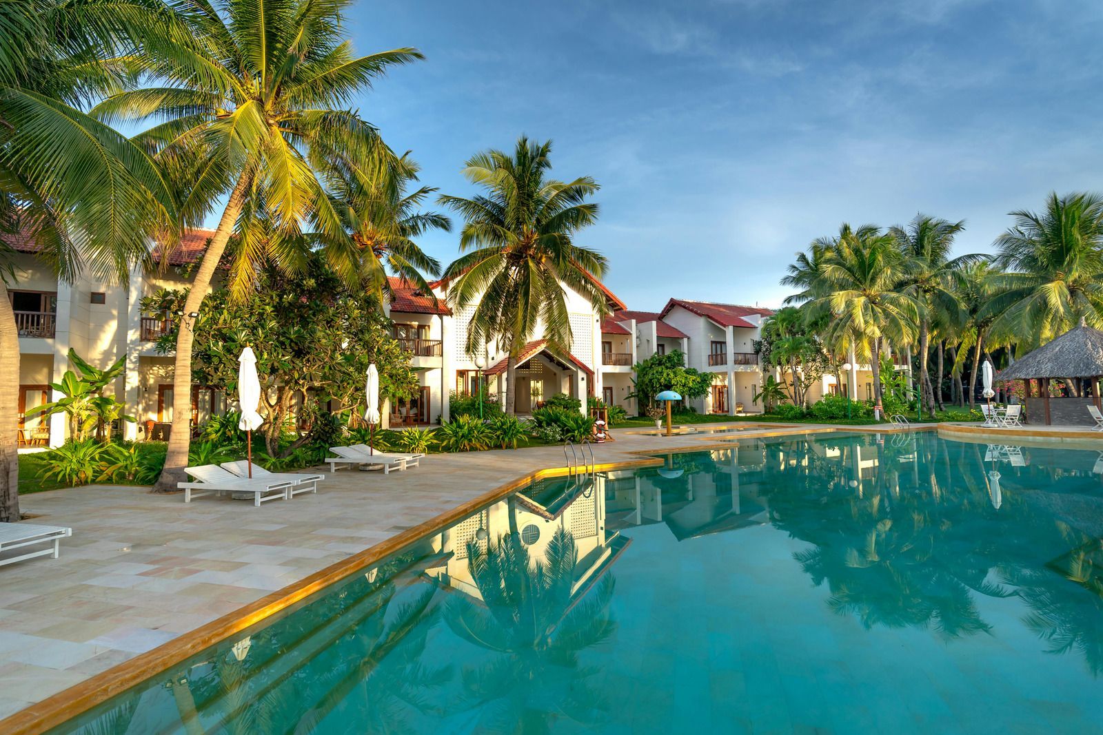 Resort pool with palm trees, buildings, and clear water reflecting the sky.