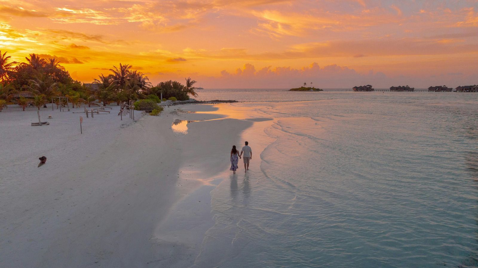 A couple walks hand-in-hand along a tropical white sand beach at sunset, with distant overwater villas under orange skies.