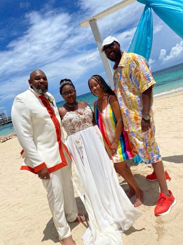 Four people on a sunny beach: a bride in white gown, groom in white suit, and two others. Blue fabric overhead.