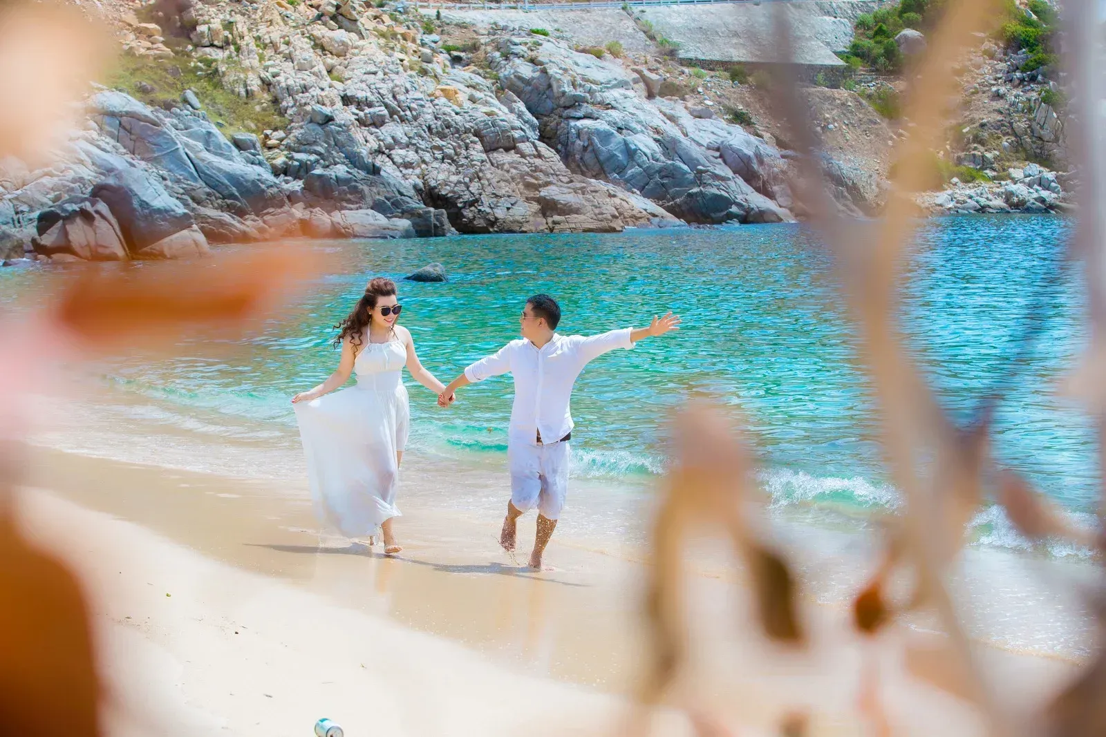 Couple holding hands, running on a beach. Woman in white dress, man in white shirt. Turquoise water, rocky shore.