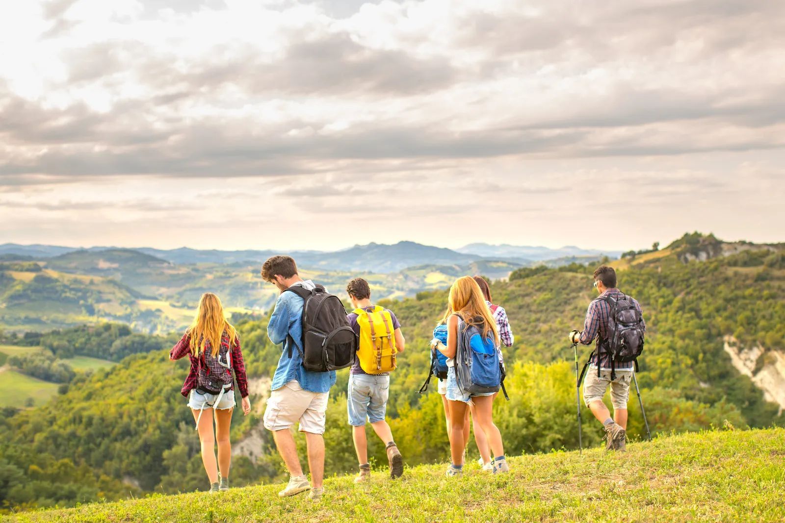 Five hikers with backpacks walking along a green hillside overlooking a valley and mountains