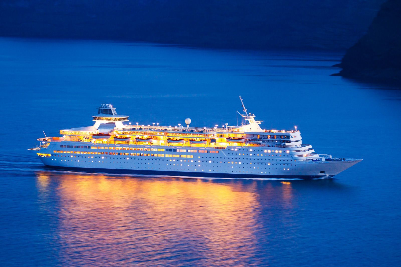 Cruise ship at night, sailing on dark blue water, with golden lights illuminating decks.