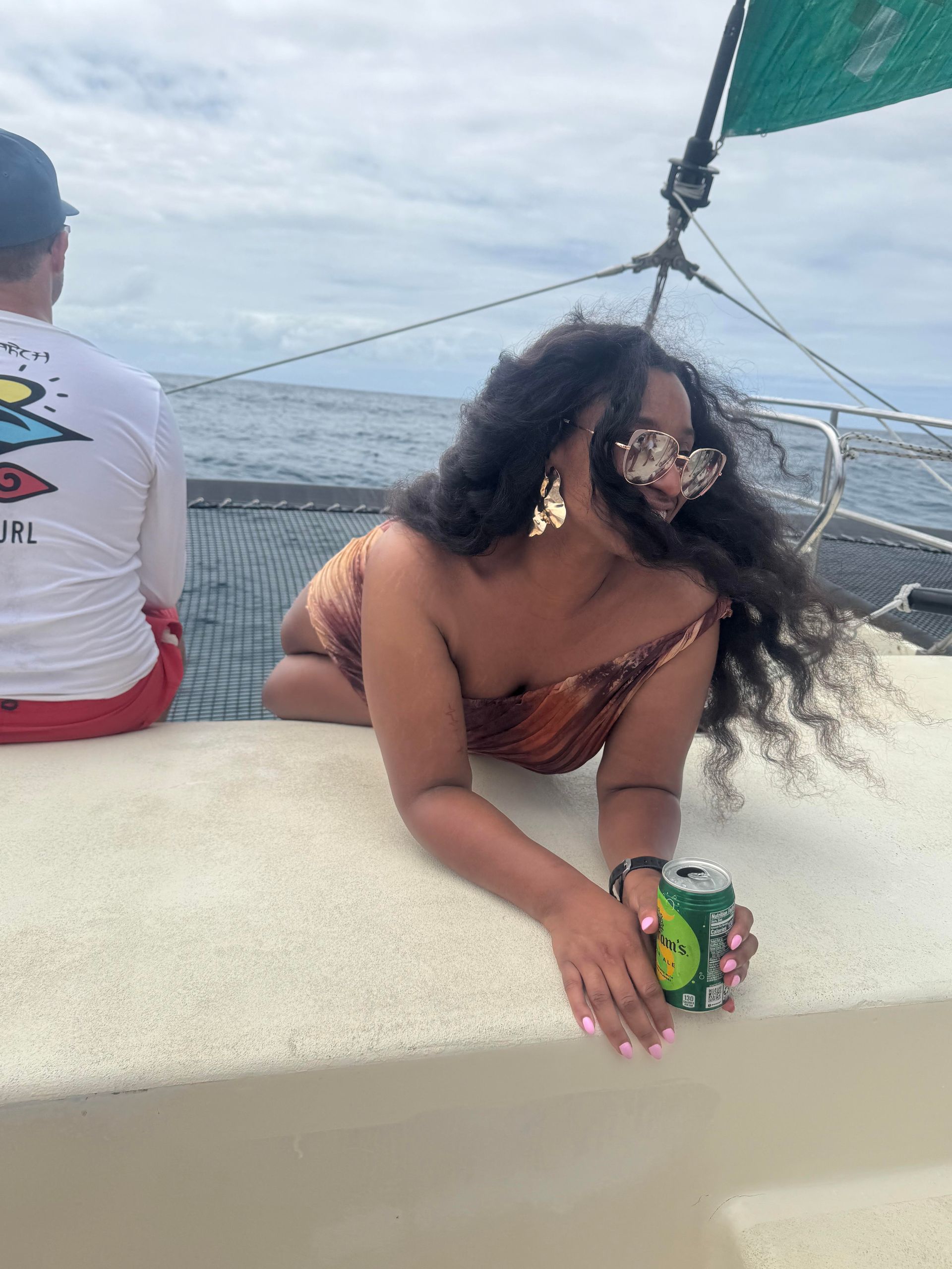 A person wearing sunglasses and a patterned top leans on a boat deck, holding a green can with the ocean in the background.