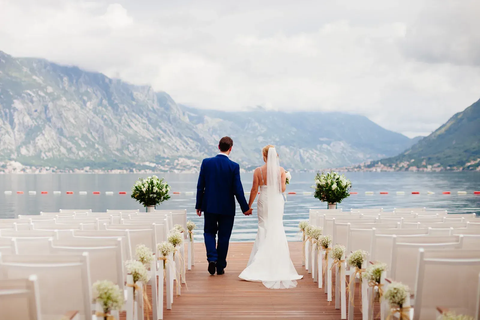 Couple holding hands, walking down a pier toward mountains, decorated for a wedding ceremony.