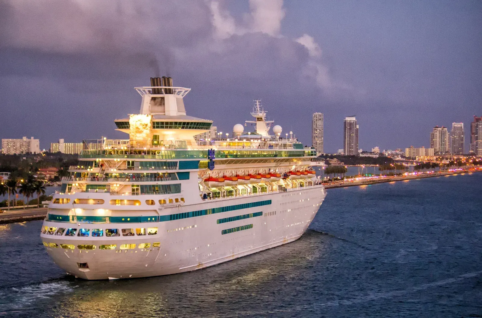 Cruise ship sailing on blue water near a city skyline at dusk; ship lights on.