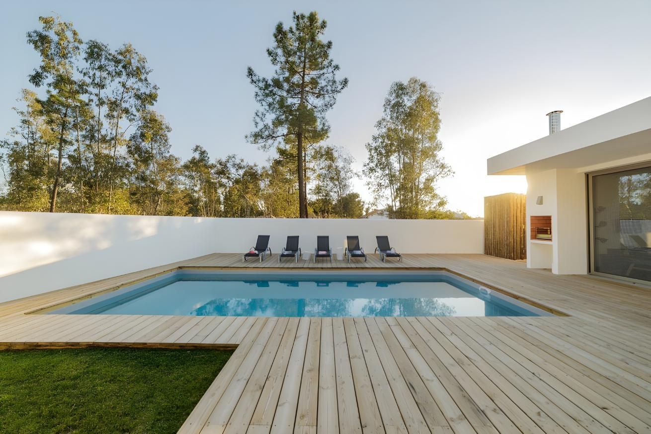 Pool With Wooden Deck and Lounge Chairs, White Wall, Trees, and a Modern White Building — Dean Thomas Pools in Manunda, QLD