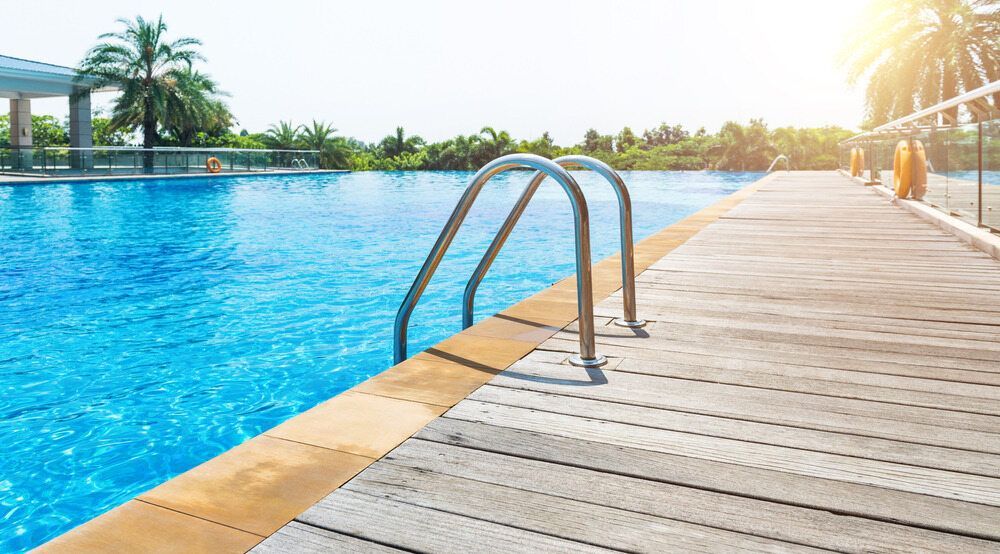 Swimming Pool With a Ladder Next to a Wooden Deck on a Sunny Day — Dean Thomas Pools in Manunda, QLD