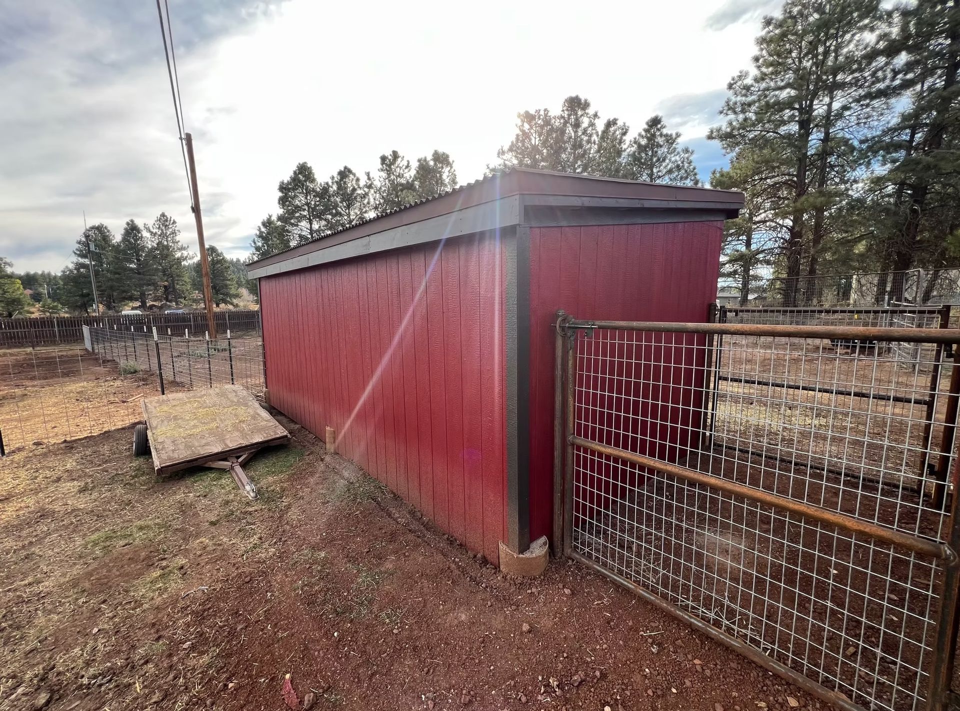 A red shed is sitting in the middle of a dirt field next to a wire fence.