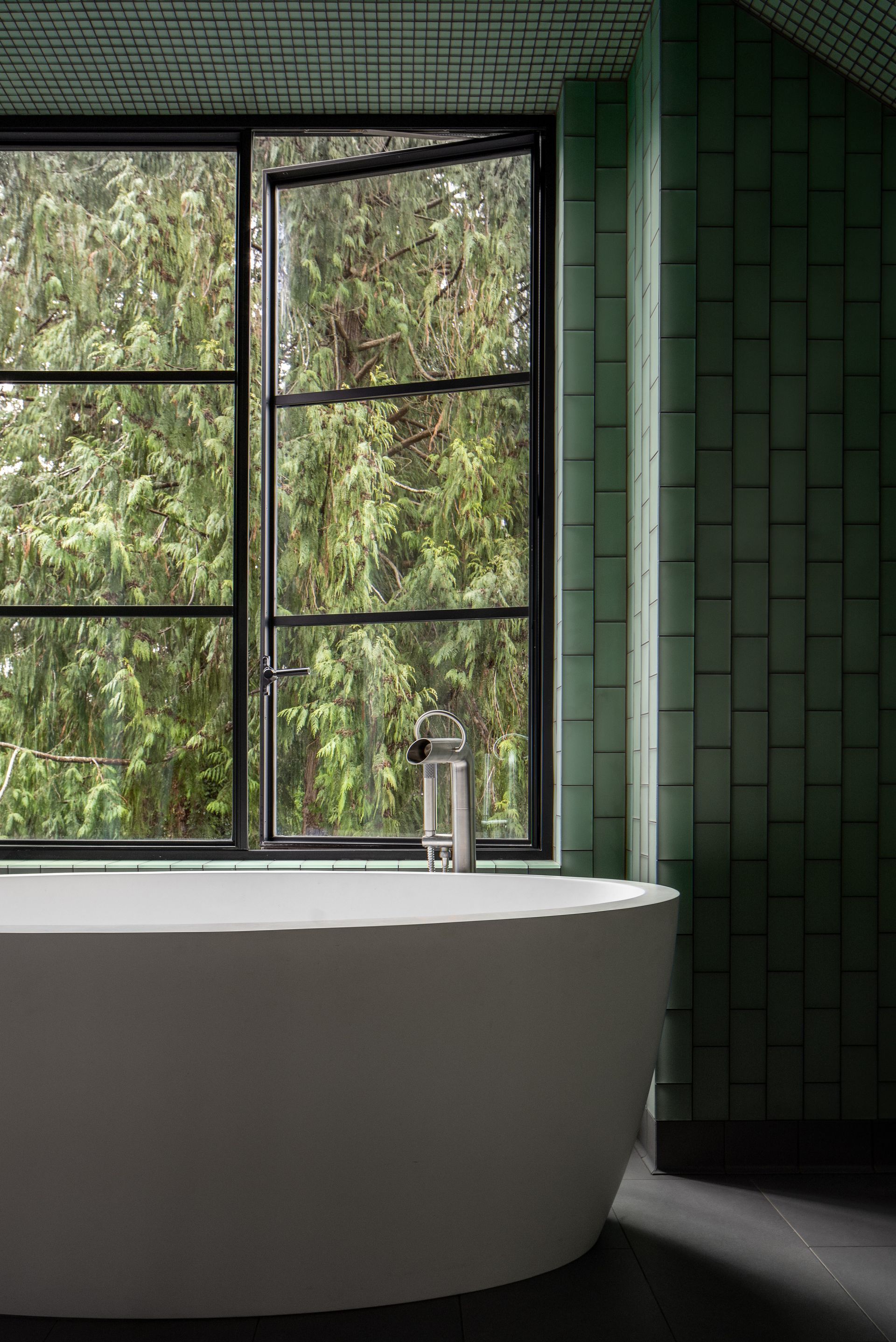 A bathtub in a room with green tiles and a large window. Bainbridge Island. Emily Barrows Photography. Wenzlau Architects.