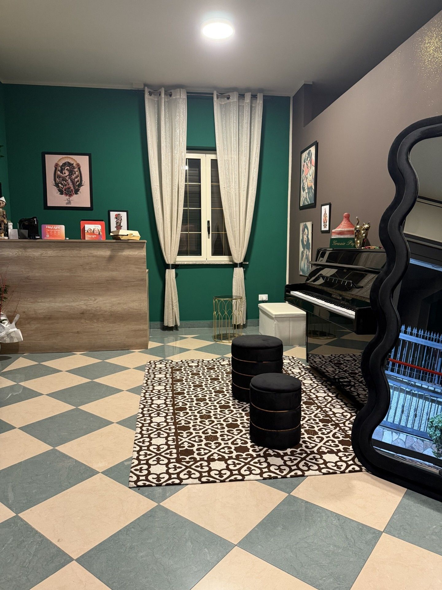 Stanza con pianoforte, pareti verdi e marroni, grande specchio, pavimento a scacchi e due sgabelli neri su un tappeto.