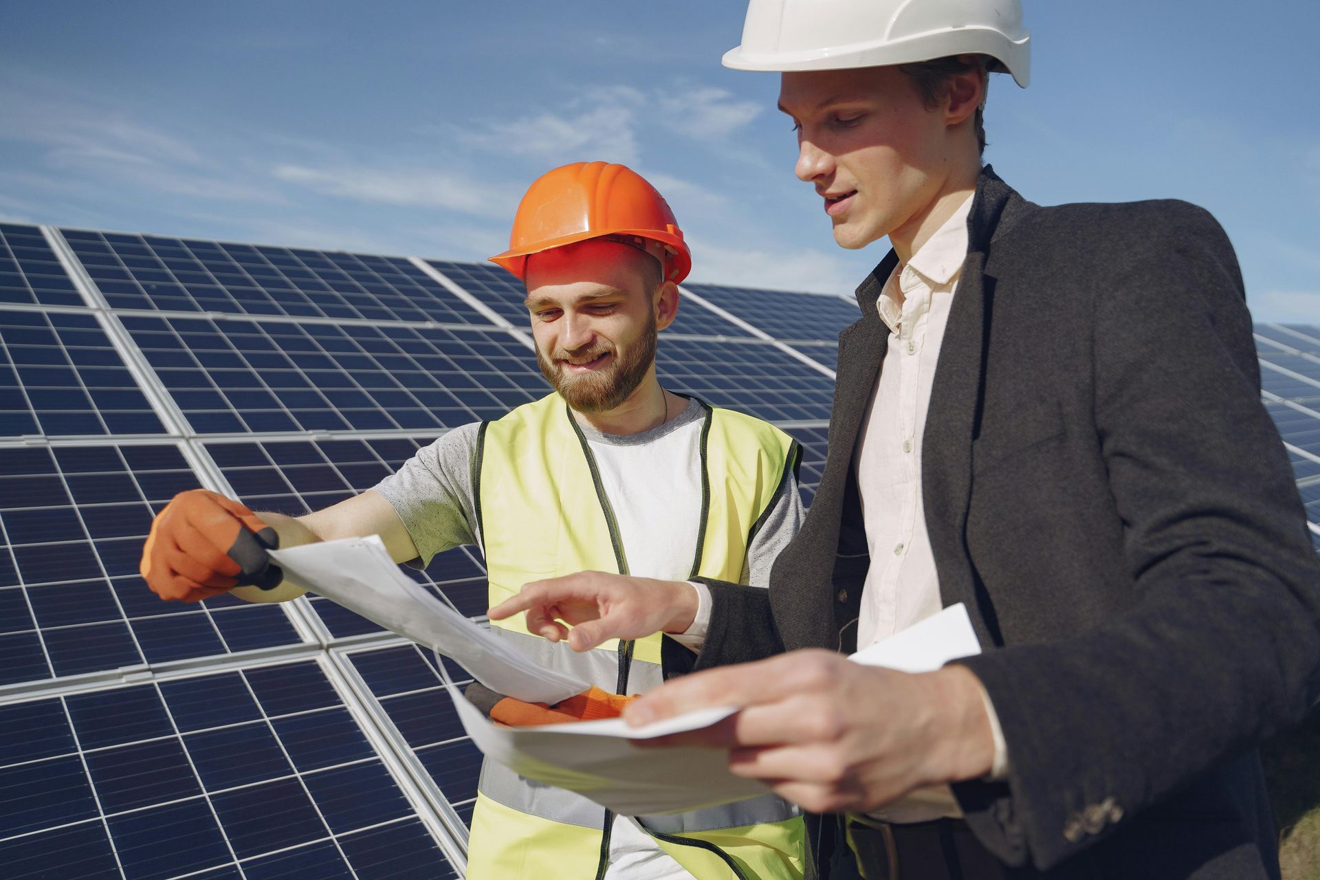 Two men reviewing blueprints near solar panels; one in a hard hat and vest, the other in a suit.