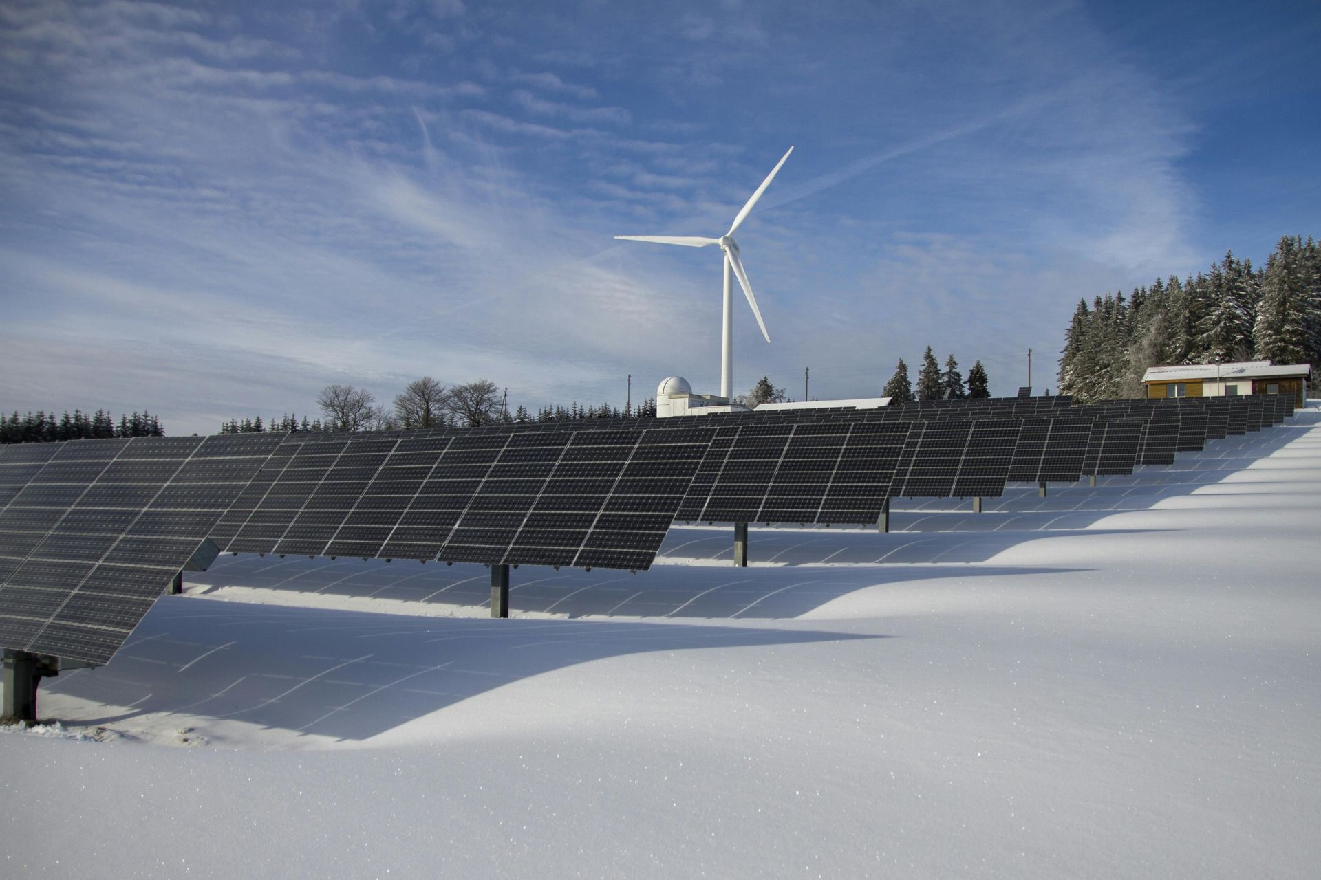 Solar panels in snow, with a wind turbine and building in the background against a blue sky.
