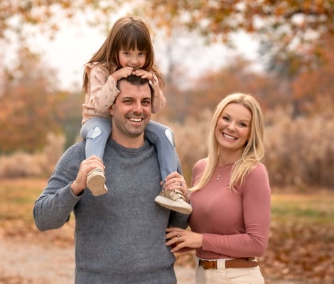 Family of three smiling outdoors: father with child on shoulders, mother beside them. Autumn foliage.
