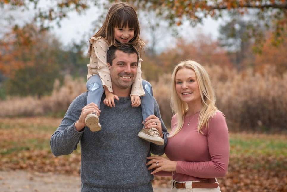 Family portrait in a garden: man holding child on shoulders; woman beside them smiling; sunny day.