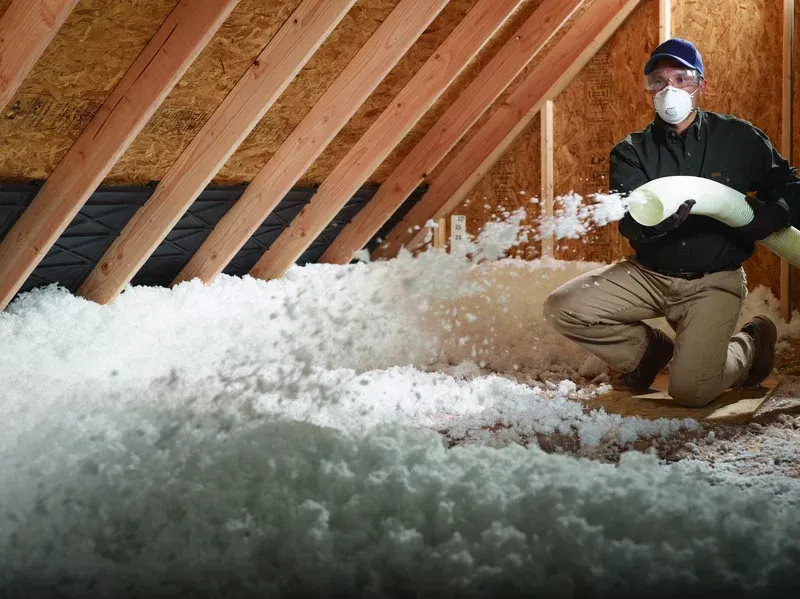 Man kneeling in an attic blowing insulation. He wears a mask and goggles, the space is wood-framed.