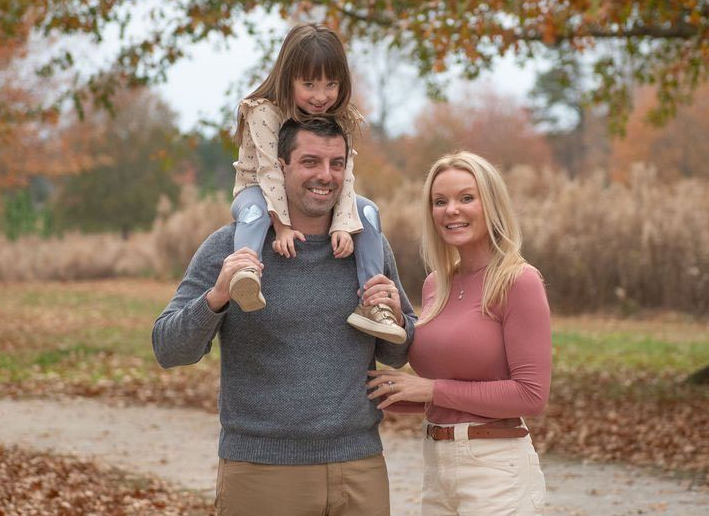Family portrait in a garden: man holding child on shoulders; woman beside them smiling; sunny day.