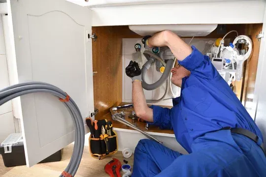 Plumber in blue overalls working under a white sink, surrounded by tools and pipes.