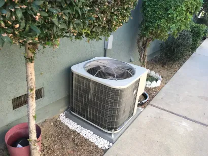 Air conditioning unit outside house, beside a tree and sidewalk, with a gravel border.