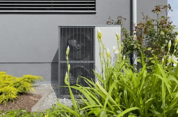 Gray heat pump unit next to building with green plants.