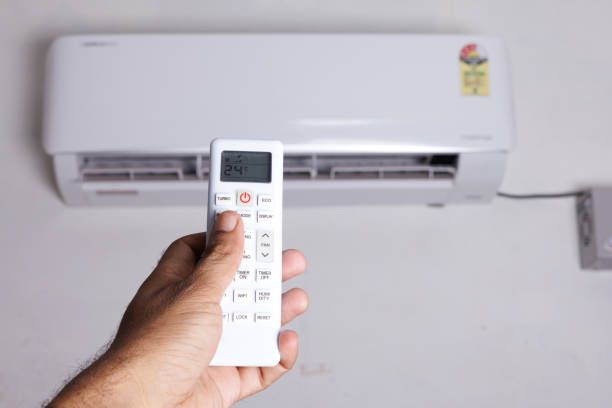Hand holding AC remote, pointing it at a white wall-mounted air conditioner.