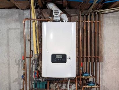 White wall-mounted boiler with digital display and copper piping in a basement setting.
