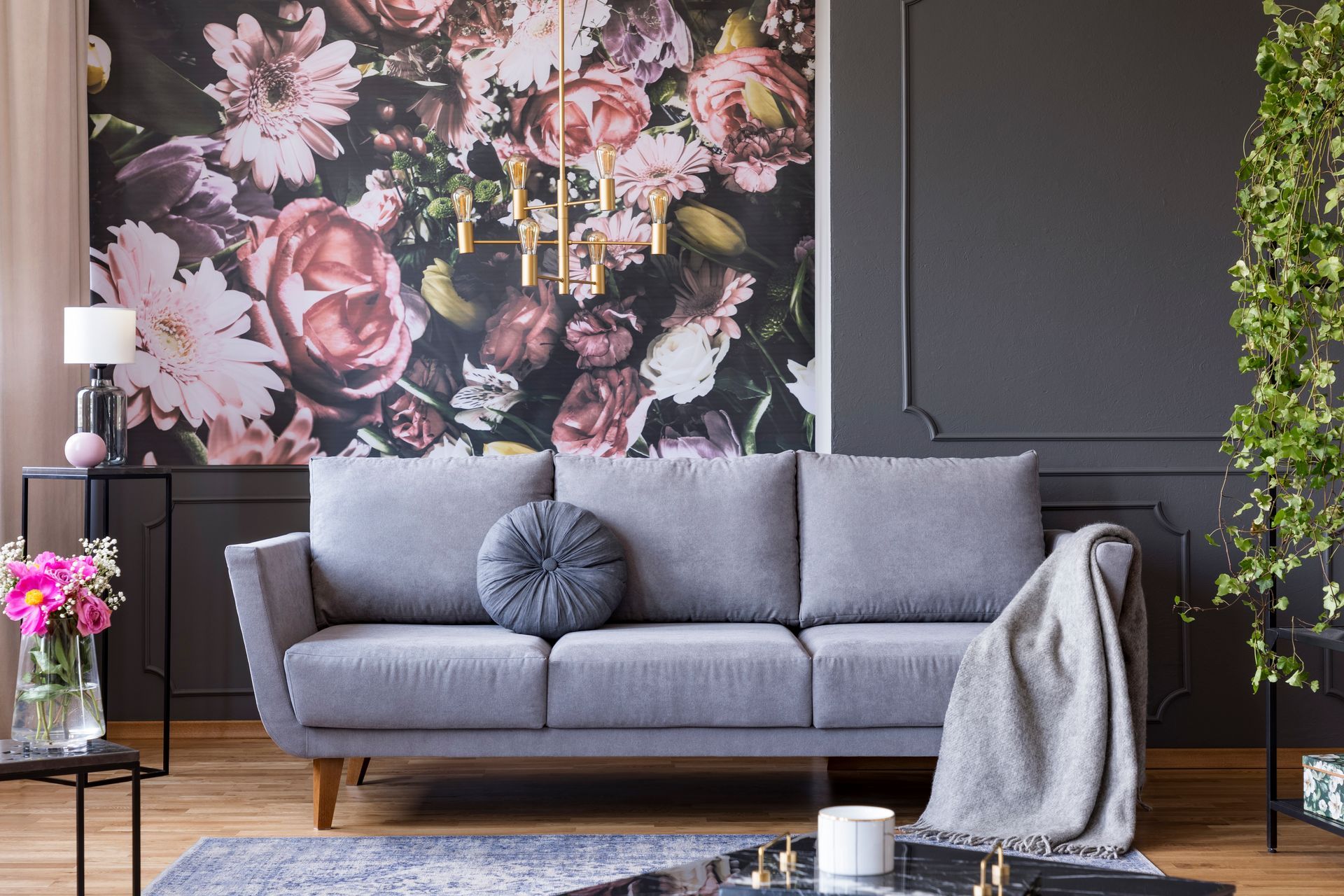A grey couch sits before a dark wall with floral wallpaper, a hanging chandelier, and a potted plant in a modern room.
