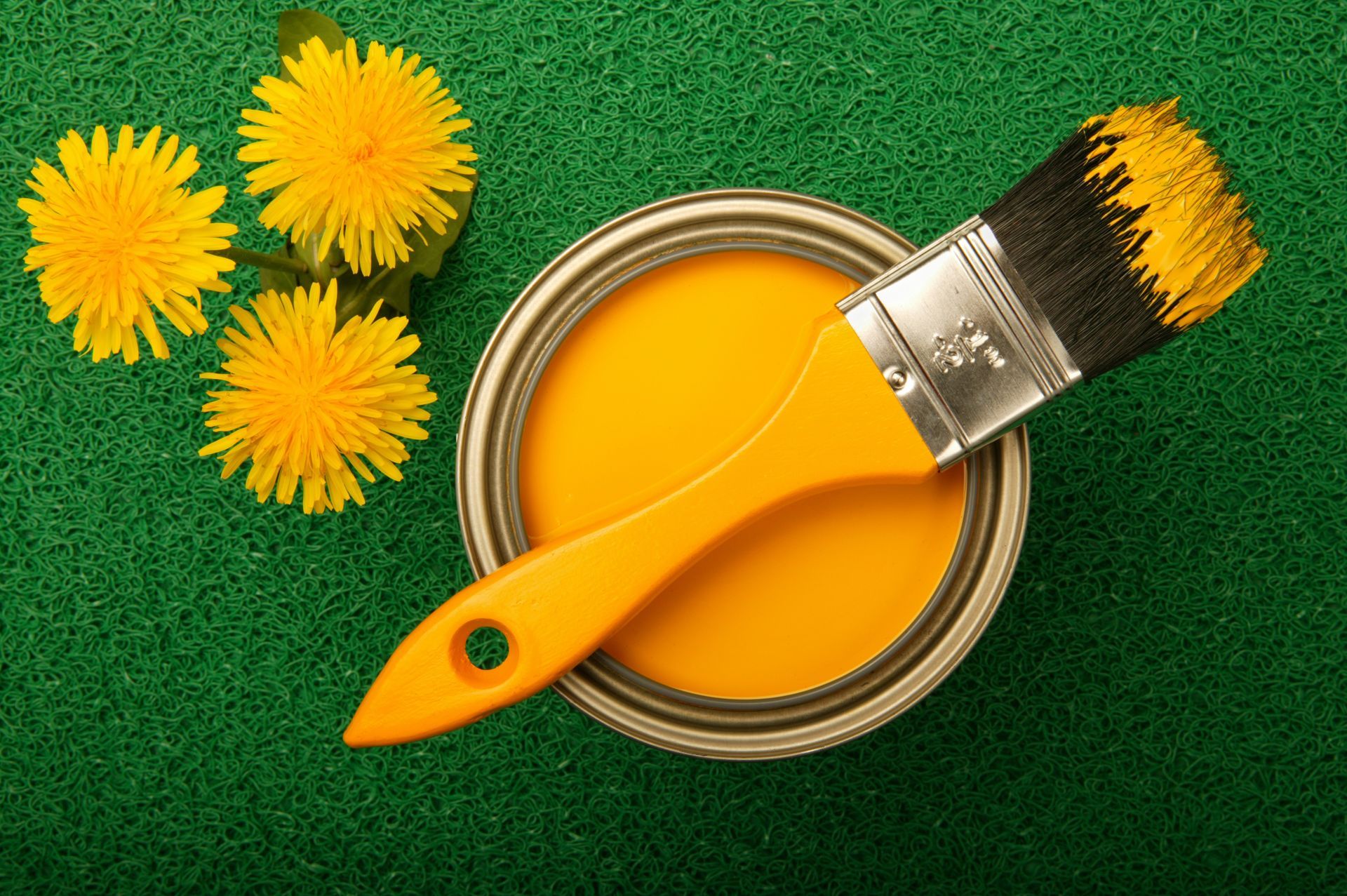 A can of yellow paint next to a brush and dandelions