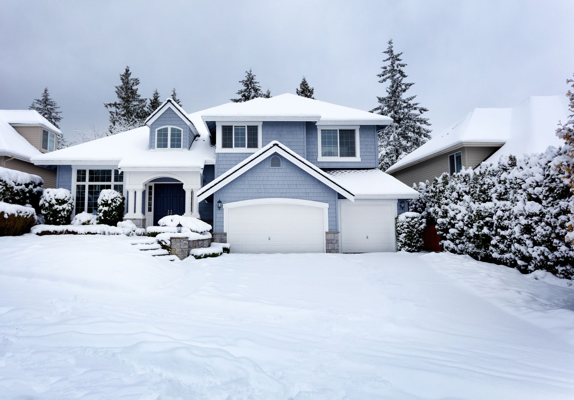 Blue house covered in snow with a two-car garage, driveway, and evergreen trees.