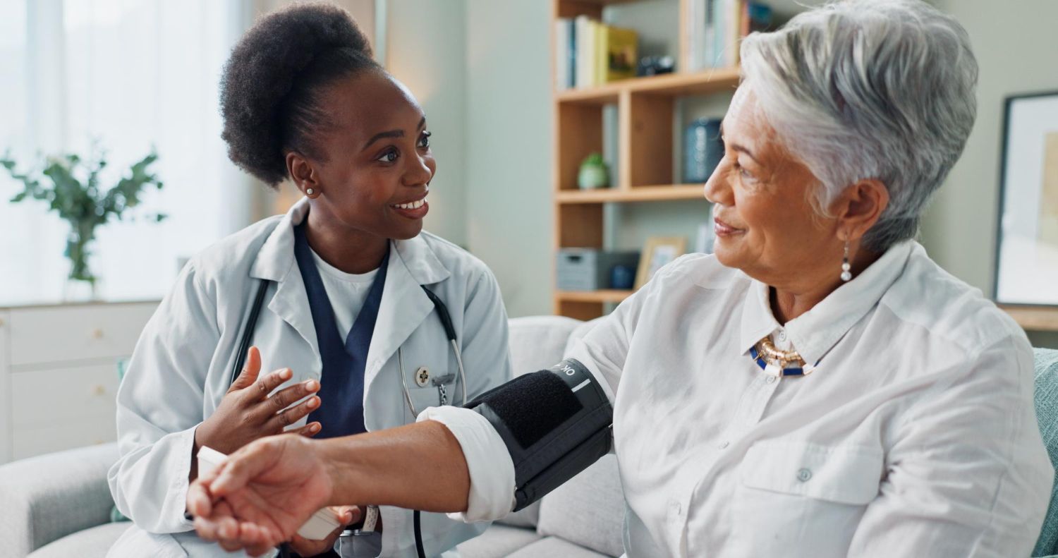 Doctor taking elderly patient's blood pressure in a home setting. The doctor is Black, and the patient is Asian.