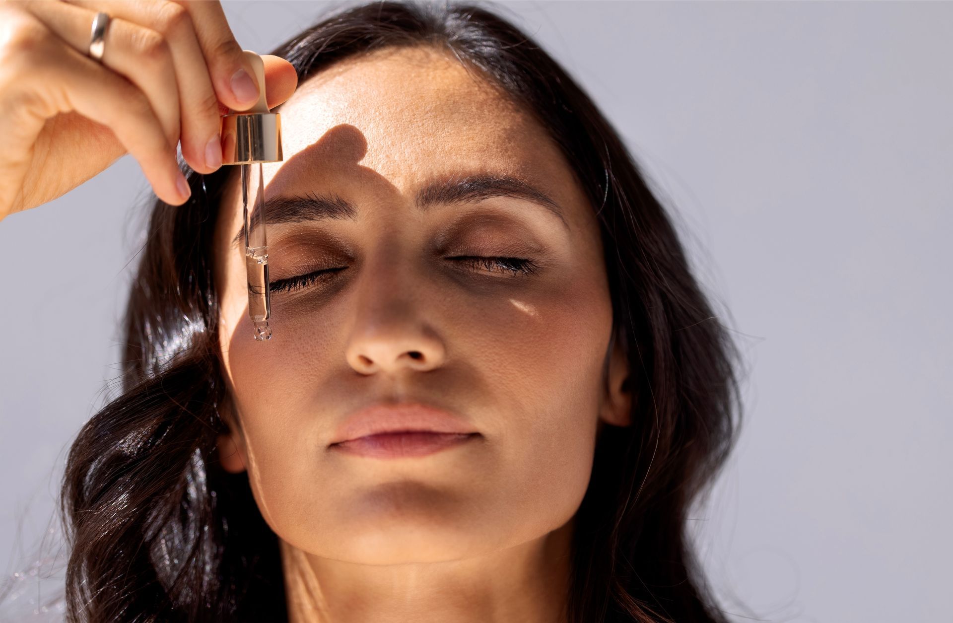 A person with closed eyes holds a glass dropper, about to apply a serum to their face under soft, natural lighting.