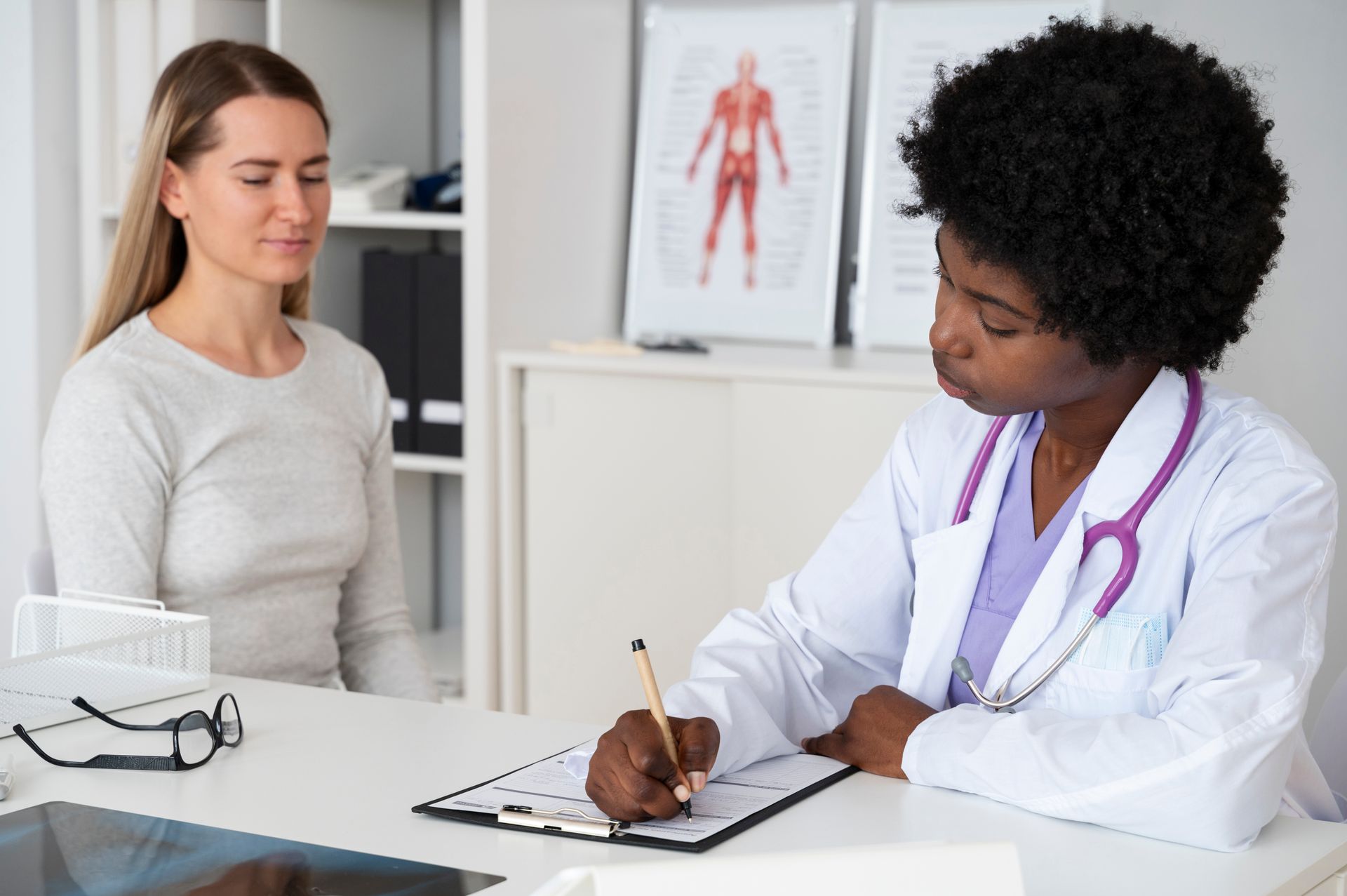 A woman is sitting at a table talking to a doctor who is writing on a clipboard.