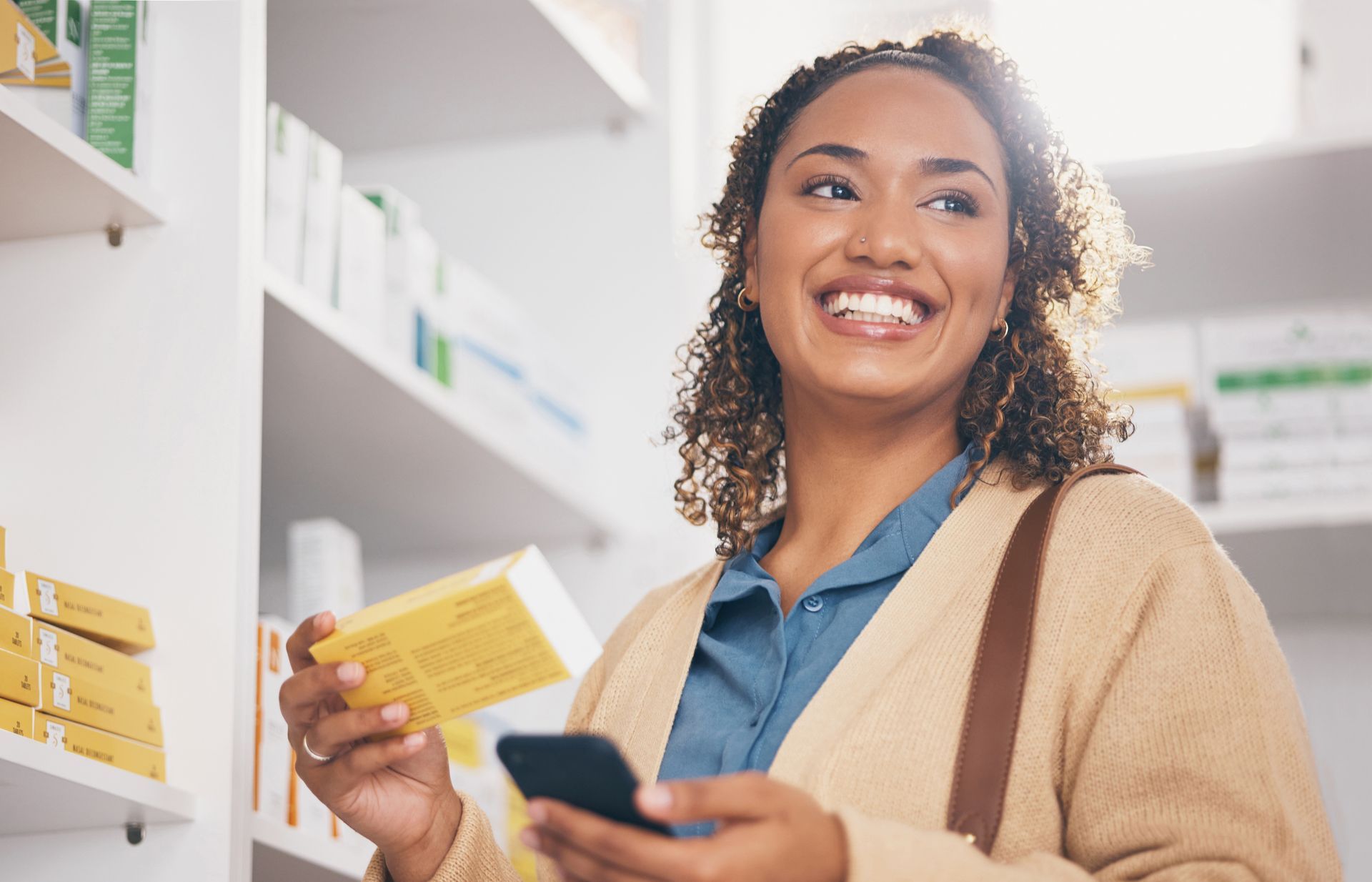 A woman is smiling while holding a box of pills and looking at her phone in a pharmacy.
