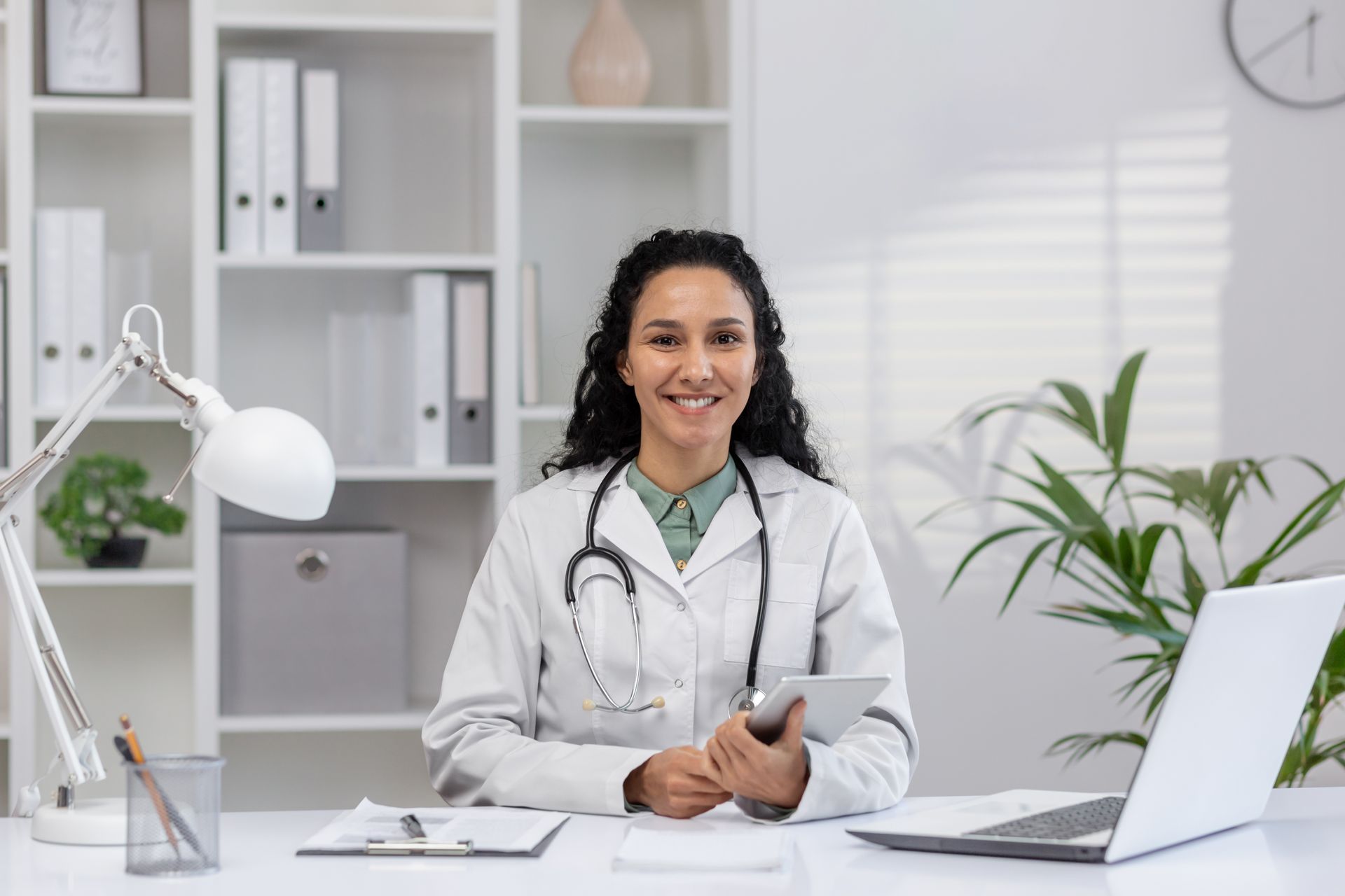 Smiling doctor in a white coat with a stethoscope, sitting at a desk with a laptop and tablet in an office setting.