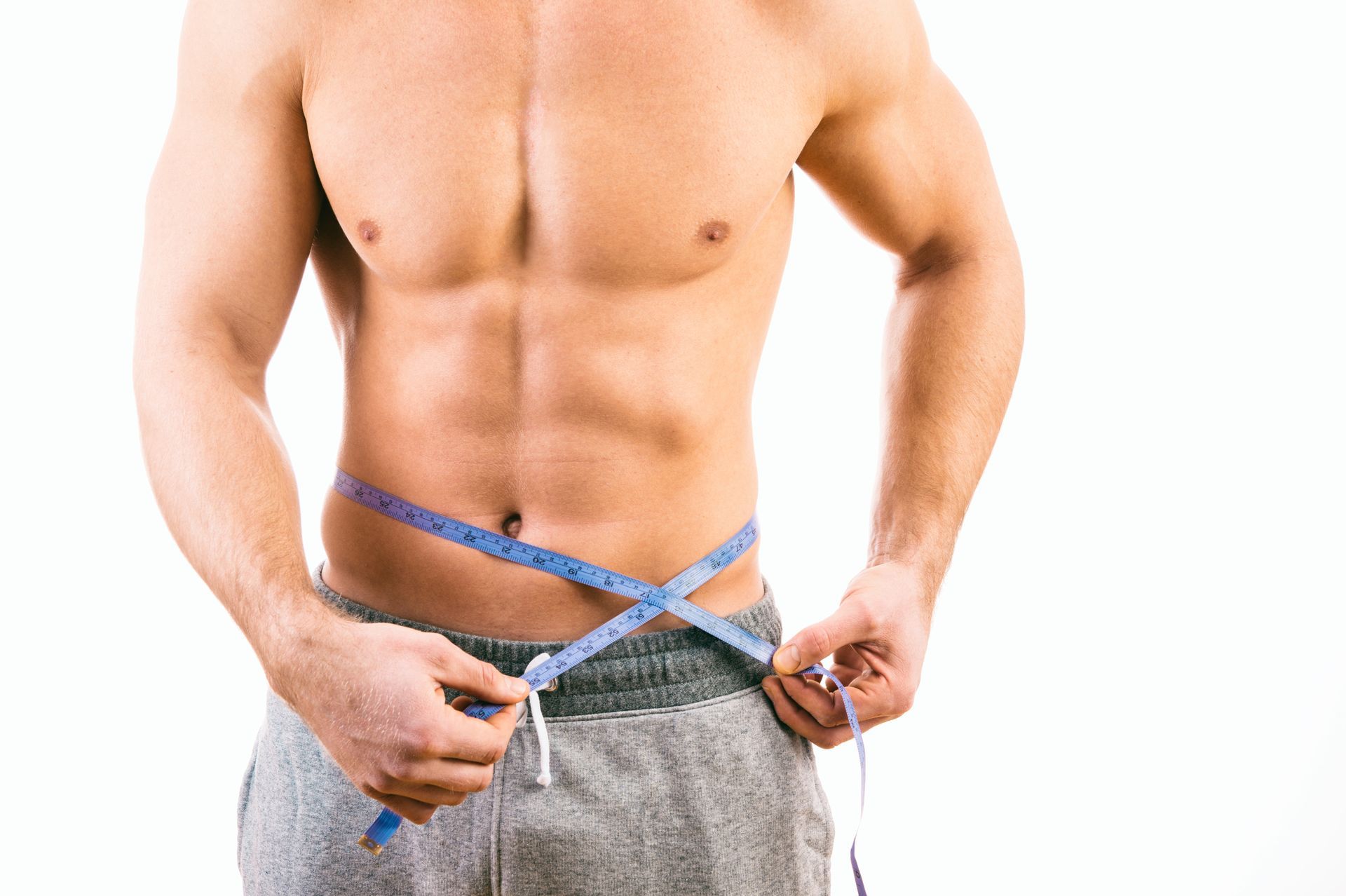 Man measuring waist with a blue tape measure. He is shirtless, wearing gray sweatpants, and has a muscular build.